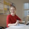 Happy woman in red sweater working at office desk with laptop and pen, smiling at camera.